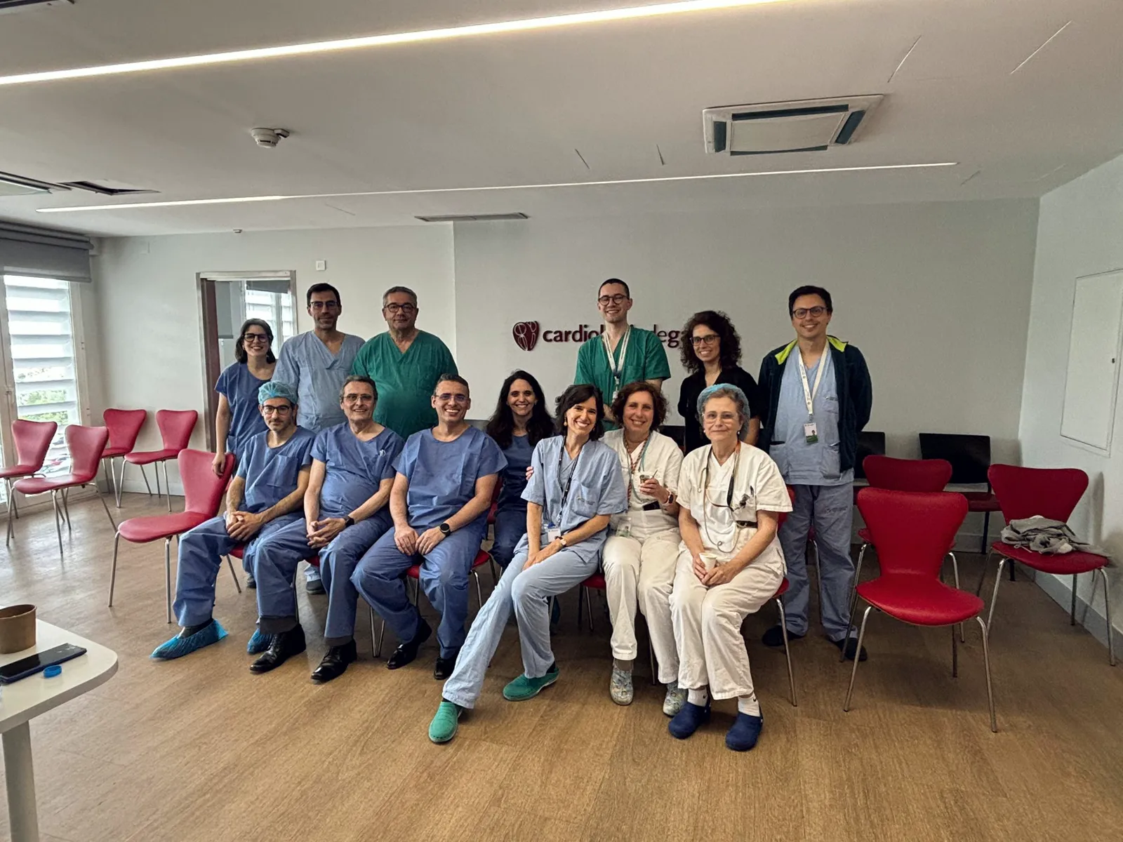 Fotografia de grupo de médicos, de pijama cirúrgico e jaleco branco, sentados e em pé em sala de reuniões hospitalar durante curso de proctoring no Centro Hospitalar de Vila Nova de Gaia, Porto, Portugal